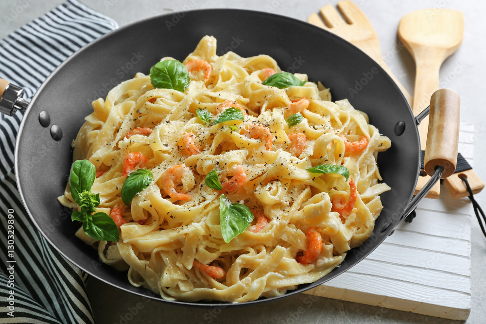 Pan with tasty alfredo pasta, kitchen board, napkin, spoon and fork on grey table