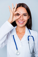 Wall Mural - Smiling female doctor  in white coat  showing OK gesture near ey