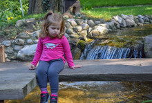 Cute Little Girl Sitting On The Old Wooden Bridge Looking At The Water In Japanese Garden On The Beautiful Sunny Day