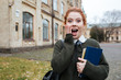 © Drobot Dean - Astonished redheaded woman student holding books outside university campus