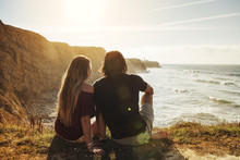 Rear View Of Couple Sitting On Cliff And Looking At Sea