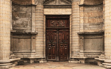 Heavy Wooden Double Door Outside Of An Old Church. Circle Wall With Square Stones And Pillars. Latin Inscriptions On The Sides And The Arch. Religious Symbolic Relief On Top. Rectangle Door Frame.