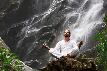 man sitting in meditation yoga on rock at waterfall in tropical