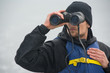 A man in a blue life jacket looking through binoculars. In the ocean