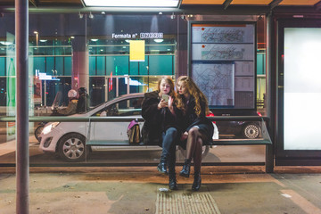 Two young beautiful caucasian women friends outdoor in the city night using smart phone hand hold sitting at bus stop - technology, social network communication concept