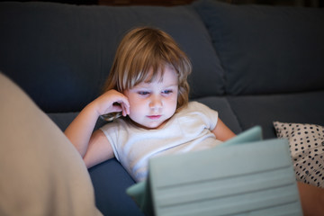 Wall Mural - blonde three years old baby shirt and shorts, sitting comfortably in sofa inside home at night reading and watching digital tablet, face illuminated by the light of the screen
