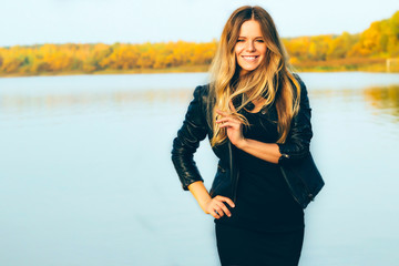 Young beautiful blonde woman in autumn park with lake in dark leather jacket smile  perfect teeth during sunset.