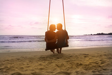 Romantic Couple Is Sitting And Kissing On Sea Beach On Rope Swing And Looking At Sunset Horizon. Family Vacation On Honeymoon