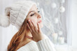 Indoor close up portrait of young beautiful girl touching her face, looking aside. Model wearing winter hat and sweater. Day light from window, white room as background. Copy space for text