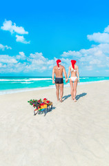 Sticker - Couple in Santa hats walking back at sea beach with sleds, Christmas tree and golden gifts