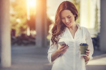 beautiful young woman is using an app in her smartphone device to send a text message in front of a sunset background