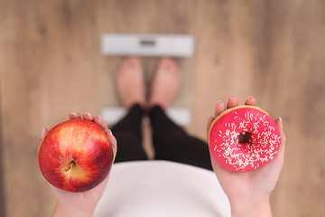 Wall Mural - Close up view of woman making choice between apple and donut with blurred scales on background. Dieting concept