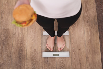 Wall Mural - Diet And Fast Food Concept. Overweight Woman Standing On Weighing Scale Holding Burger ( Hamburger ). Unhealthy Junk Food. Dieting, Lifestyle. Weight Loss. Obesity. Top View