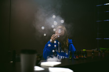 Young Cute Woman, The Seller In Vape Shop Shop, Smoking Electronic Cigarette,  Vaping And Releases A Cloud Of Vapor And Inflates Soap Bubbles