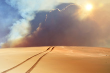 Vehicle Tracks Through Desert And Dunes Leading Into A Sand, Smoke And Cloud Filled Sky. 