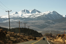 View Of A Straight Section Of Route 395, With Arid Desert On The Road-side, A Line Of Electric Wires Poles And Snowy Mountains To Close The Horizon Against A Clear Blue Sky