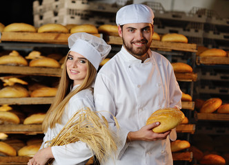 Girl with a guy in white overalls are holding fresh bread and bakery in the background of shelves with bakery