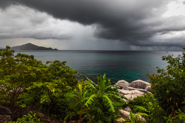 Canvas Print - Dark rainy clouds over tropical sea with stones and plant on the foreground