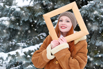 beautiful woman portrait on winter outdoor, look through wooden frame, snowy fir trees in forest, long red hair, wearing a sheepskin coat