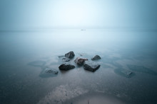 Coastal Landscape Of Foggy River With Fisherman In The Boat As Small Detail. Stones In The Water On Foreground. Long Exposure Shot.