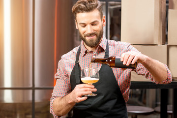 Handsome brewer in apron and shirt pouring beer into the glass at the manufacturing