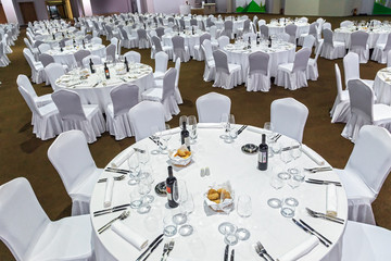 Plenty of big round tables and chairs covered with white tablecloth set for a meal in a restaurant. Wide angle interior view