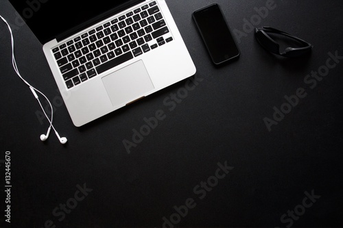 Fotografie, Obraz Laptop, smart phone, sunglasses and earbuds on dark, black desk