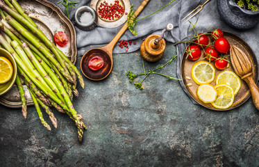 Wall Mural - Tasty asparagus cooking with tomatoes, lemon and seasoning, preparation on rustic kitchen table with wooden spoon, top view, border. Dark style