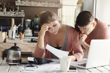 Young married couple facing financial problem during economic crisis. Frustrated woman and unhappy man studying utility bill in kitchen, shocked with amount to be paid for gas and electricity