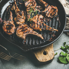 Barbecue dinner. Close-up of grilled lamb meat chops with onion and rosemary in cast iron pan on wooden board over grey background, selective focus, square crop. Slow food concept