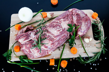 Big piece of raw beef meat with vegetables onion, carrot, garlic and spices pepper, rosemary, parsley, sea salt on a wooden cutting board.
