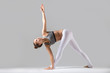 © fizkes - Young attractive woman practicing yoga, standing in Revolved Triangle exercise, Parivrrta Trikonasana pose, working out wearing sportswear, indoor full length, isolated against grey studio background