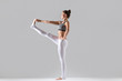 © fizkes - Young attractive woman practicing yoga, standing in Extended Hand to Big Toe exercise, Utthita Hasta Padangustasana pose, working out wearing sportswear, indoor full length, grey studio, profile