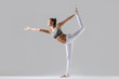 © fizkes - Young attractive woman practicing yoga, standing in Lord of the Dance exercise, Natarajasana pose, working out wearing sportswear, white pants, gray top, indoor full length, grey studio background