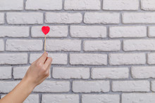Woman's Hand Holding Red Heart Shape On Stick. White Brick Wall Baskground. Valentine Concept.