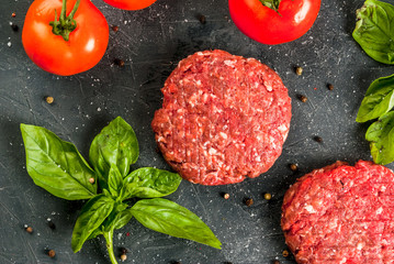 Fresh raw home-made minced beef steak burger with spices, basil and tomatoes on a cutting board on a stone table, copy space, top view