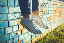 Female Legs In White Canvas Sneakers And Jeans Dangling From A Blue Brick Wall On A Sunny Summer Day