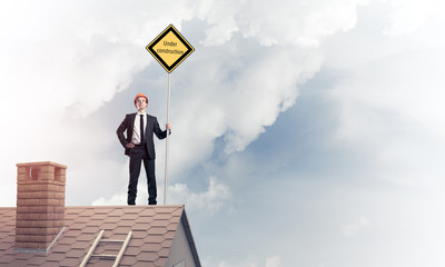 Young businessman on house brick roof holding yellow signboard. 