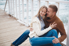 Young Couple Blond Guy With Short Hair In A Brown Sweater And Blue Jeans And A Blonde Girl With Straight Long Hair,spend Time Together,sitting,embraced On A White Wooden Pier Near The Sea In Autumn