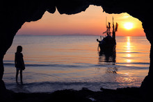 Girls Stands In Front Of The Cave By The Sea With Local Fishing Boats And Sunset Background. 
