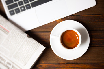 Cup of espresso with laptop and newspaper on wooden desk, top view