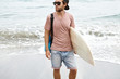 Attractive young surfer wearing trendy shades and snapback holding surfboard under his arm, standing on wet sandy beach, waiting for his friends to join him in their morning surfing exercise