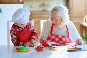 Happy family, grandmother with her granddaughter, preparing delicious pizza together topping it with tomato sauce, vegetables and cheese, sitting at white dining table at bright sunny room at home