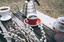 Fresh Morning Coffee In A Red Cup On The Old Wooden Table With A Kettle, Coffee Dripper And Some Pussy Willow Outdoor Picnic