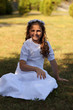 Śliczna dziewczynka w białej sukni siedzi na trawie.