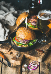 Wall Mural - Homemade beef burgers with crispy bacon and vegetables in small pan and glass of wheat beer on rustic serving board over shabby wooden background, selective focus, vertical composition