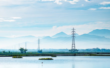 Transmission Tower, Electricity Pylon Or A Power Pylon, Standing Majestically Lined Up To The Horizon, In Front Of Large Lake Or Pond, Also With Beautiful Cloud And Mountain In The Background, Capture