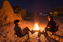 Couple Sitting At Burning Camp Fire In The Night. Camping In The Desert With Wild Elephants In Background. Summer Adventures And Exploration In African National Parks. Camping Stove And Gas Burner.
