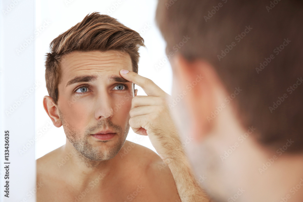 Young man standing in front of mirror and touching his face