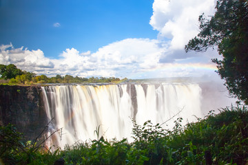 Wall Mural - Victoria Falls.  Frontal view with a rainbow.  Taken with an MD filter.  Blue sky with clouds looming.